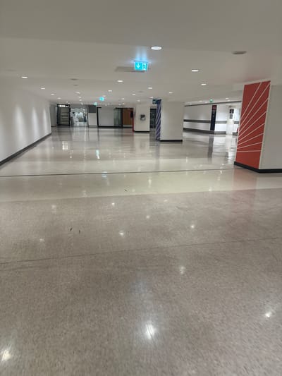 The image displays an empty, modern interior hallway, likely within a public building such as an airport terminal in Paray-Vieille-Poste, France. The perspective looks down a long corridor, which is devoid of people.

The ceiling, a smooth white surface, features numerous evenly spaced recessed spotlights, casting bright, round reflections on the highly polished, speckled grey floor. Several green emergency exit signs with white arrows and human figures are suspended from the ceiling, indicating egress routes to the viewer's left.

The walls are predominantly white, punctuated by dark trim lines along the floor and ceiling. Further down the corridor, on the right wall, there's a distinctive section with a bold red and white striped pattern, resembling a sunburst or radiating lines. Several black and white directional signs are visible on the walls in the distance, though their text is largely illegible due to the distance. One sign near the middle of the corridor appears to include a pictogram of a person and stairs, possibly indicating a staircase or elevator.

The floor is a light grey, speckled material, possibly terrazzo or polished concrete, and is highly reflective, mirroring the ceiling lights and the structural elements of the corridor. The overall atmosphere is clean, quiet, and contemporary, illuminated solely by artificial light, making the time of day indeterminate.