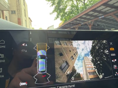 The image shows a car's infotainment screen displaying the parking assist system with a 360-degree view. The display shows a stylized top-down view of the vehicle surrounded by proximity sensors. The screen also shows the live camera feed from the surroundings. The buildings visible in the camera feed are located in Târgu Mureș, Romania. The buildings are located on Strada Bolyai and Strada Mihai Viteazul. The image also includes elements of the car interior and the outside environment, including a roof structure and trees.