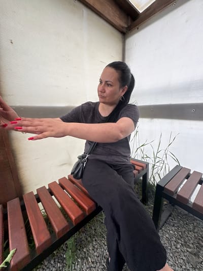 A woman with dark hair pulled into a ponytail sits on a wooden bench in what appears to be an outdoor covered area, possibly a greenhouse or a sunroom. The walls of the structure are made of corrugated translucent material, and wooden beams support the ceiling. The woman is wearing a dark grey t-shirt and black pants. She has long, bright red acrylic nails and wears a simple hoop earring in her left ear. A small black crossbody bag is slung over her right shoulder. Her left arm is extended forward, with her hand open, as if reaching out or presenting something. Another bench, similar in design with wooden slats, is visible to her right, and the ground is covered in small grey stones and some sparse green vegetation. The lighting suggests it is daytime. The location is identified as Zaliznychne, Ukraine.