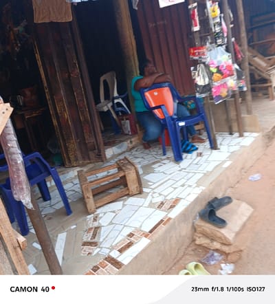 A woman in a green shirt and blue jeans is seated in a blue and orange plastic chair outside a shop. She appears to be resting, with her arms crossed on the back of the chair. The shopfront is weathered and has wooden and metal panels. Other plastic chairs and a small wooden stool are nearby. Assorted merchandise hangs for sale in front of the shop. The ground is a mix of tiled flooring and dirt. A pair of black sandals rests on a stone.