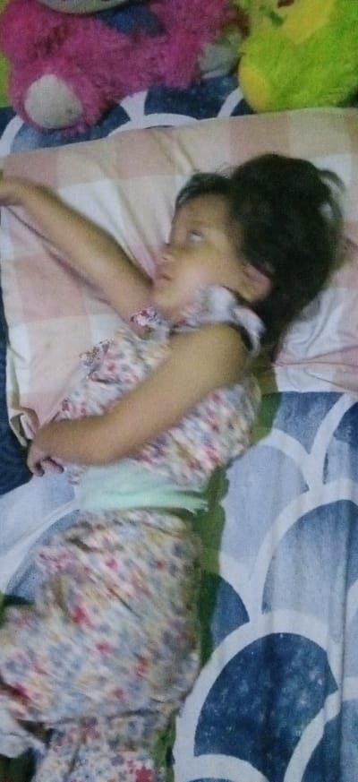 The image captures a young child lying on a bed, appearing to be asleep or resting. The child is positioned on their back, with their head turned slightly to the left, eyes closed or mostly closed, and one arm extended upwards over a pillow. They are wearing a light-colored, floral-patterned dress or sleepwear with a light green band around the waist. Their dark hair is spread around their head on the pillow.
The setting appears to be indoors, likely a bedroom. The child is on a bed covered with a blue and white patterned sheet. A light-colored pillow with subtle striped patterns (pink, white, and tan) supports the child's head. In the background, two stuffed animals are visible: a prominent pink teddy bear and a less distinct green stuffed animal, suggesting a child's sleeping area.
The overall impression is one of peaceful rest. Due to the indoor setting, the time of day and weather cannot be determined from the visual information alone. There are no specific visual cues within the image to indicate the provided location of Banjaran, Indonesia. The image is slightly blurry, particularly around the child's face and arms. There is no visible text in the media file.