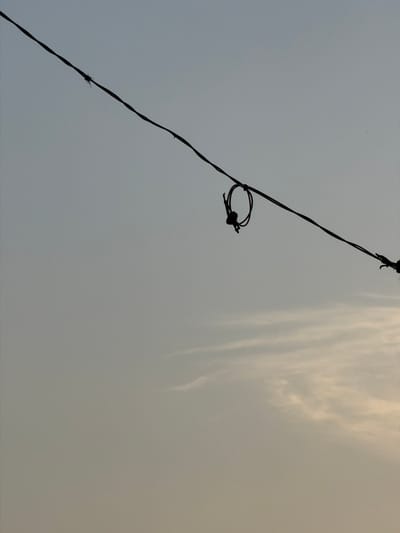 The image depicts a wire strung diagonally across a pale, overcast sky. A small, dark, looped object, possibly a knot or a piece of debris, is attached to the wire in the center of the frame. A very faint wispy cloud formation is visible in the lower portion of the sky. No people or structures are visible. The location is identified as Grao, Spain.