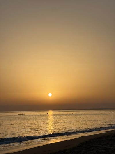 This image displays a sunrise over the Atlantic Ocean, captured from a beach in the vicinity of Almancil, Loulé, Portugal. The sky, occupying approximately the upper two-thirds of the frame, exhibits a gradient of color, transitioning from a bright golden-orange at the horizon to a muted, lighter orange-brown in the upper portion. The sun, a distinct, intensely bright white-yellow disc, is positioned low on the horizon, slightly to the left of the image's vertical center. A prominent vertical column of sunlight reflection extends from the sun across the calm ocean surface towards the foreground. The ocean, which fills the mid-ground, shows minimal wave activity. A dark, rectangular floating platform is situated in the water in the middle-left, and several dark, spherical buoys are visible at varying distances across the water. The foreground features a dark sand beach, with small, white-crested waves breaking gently along the shoreline. No individuals, active subjects, or direct interactions are visible within the scene.