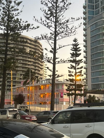 This photograph depicts an urban street scene in Coolangatta, Australia, during either sunrise or sunset. The sky is a soft, diffused gray, with a bright, golden orb of the sun peeking through the branches of a tree on the right side, casting a warm glow.
In the immediate foreground, several tall, slender trees, likely Norfolk Pines, frame the view. Their dark, needle-like branches are silhouetted against the sky, some appearing to be swaying slightly, suggesting a gentle breeze.
Behind the trees, the scene unfolds with a mix of architectural styles. To the left, two modern, multi-story buildings dominate the skyline. One features a distinctive wavy, curvilinear design with numerous balconies, while the other is a more rectilinear structure with many windows. Further to the right and slightly below these high-rises, a multi-story building complex, predominantly pink or peach-colored, stands out with its warm interior lights glowing and decorative string lights draped across its facade, suggesting a festive or welcoming atmosphere. This building has visible external staircases and an open design, possibly a motel or apartment complex.
To the far right, several vehicles are parked along the street, partially obscuring the view of the buildings. A white van or SUV is most prominent, with a portion of a red car visible behind it. In the distant background, on a white sign above the pink building and behind the parked cars, the text "COOLANGATTA" is clearly visible, confirming the location. The overall impression is one of a vibrant coastal town transitioning from day to night or vice versa, with a blend of contemporary and possibly retro-styled architecture.
