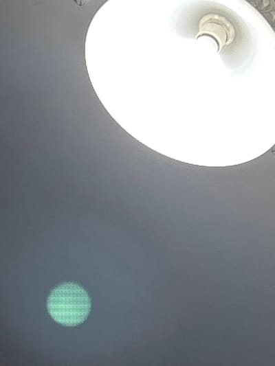 The image shows a ceiling light fixture and a green circular artifact on a dark gray surface. The light fixture is white with a metal socket for a bulb. The green artifact appears to be a reflection or lens flare, with a grid-like pattern visible within it. The context of Lyaskovets, Bulgaria is not visually represented in the image itself.