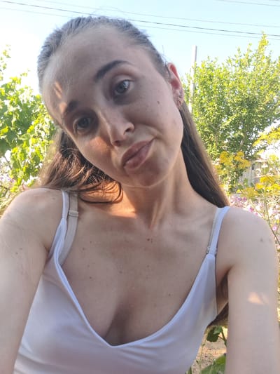 A woman is pictured in a selfie, taken outdoors in Vypasne, Ukraine. She is wearing a white tank top that reveals her chest and shoulder. Her hair is dark brown and long, falling over her shoulder. The woman's face is close to the camera, and she is looking directly at it with a neutral expression. She has brown eyes and light skin with some freckles. A silver earring is visible in her left ear.

Behind the woman, lush green foliage and some yellow flowers are visible, suggesting a natural, possibly rural, setting. The sky is a clear blue, indicating a sunny day. The lighting suggests it is daytime, likely late morning or early afternoon, given the angle of the sun and the shadows. The overall mood of the image is casual and relaxed. There is no visible text in the image.