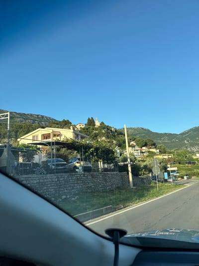 The image captures a view from inside a vehicle, looking out towards a hillside residential area in Kaluđerac, Montenegro. The sky is clear and blue. A light yellow house with a balcony and air conditioning units is prominently visible on the hillside, surrounded by trees and other smaller buildings. Several vehicles, including cars and a motorcycle, are parked in front of the house behind a stone wall. A paved road runs alongside the property. In the distance, a white structure, possibly a church, is visible atop a hill, with more houses scattered across the slopes. The overall environment is a sunny, suburban setting with a mountainous backdrop.