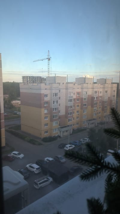 The image captures a view of a residential building under construction, likely in the city of Kovrov, Russia, given the context. A large yellow and brick-colored apartment building dominates the center of the frame, featuring numerous windows and air conditioning units. In the background, a construction crane is visible against a clear sky. A street with parked cars, including several white vehicles, a blue sedan, and a dark SUV, runs alongside the building. A partially constructed building is also present in the distance, indicating ongoing development in the area. The foreground is partially obscured by the branches of a coniferous tree, suggesting the photo was taken from a window. The overall scene depicts an urban environment during daylight, with a focus on residential construction and infrastructure.