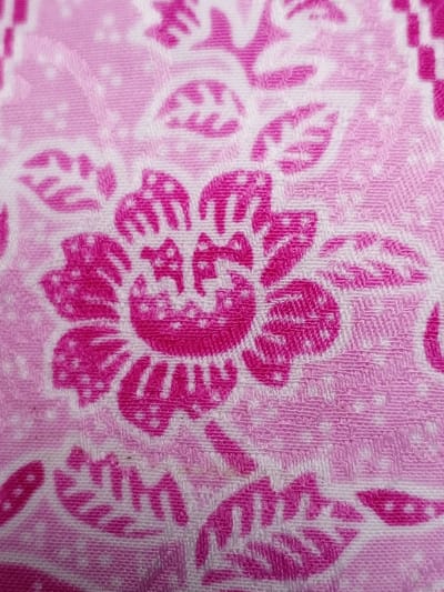 The image showcases a close-up of fabric with a floral pattern. Predominantly pink and white, the design features stylized flowers with intricate petal details and surrounding leaf motifs. The fabric's texture is evident, with a subtle interwoven pattern visible beneath the floral print. The color palette ranges from a light pastel pink to a vibrant magenta, creating visual depth and contrast against the white background. The image appears to be a macro shot, emphasizing the details of the fabric and the floral design.