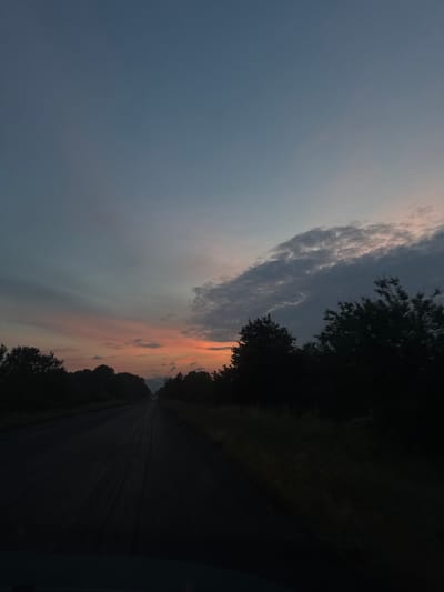 The image captures a serene, dusky scene along a road, likely taken from a moving vehicle. The primary subject is a vast sky showcasing a sunset, accompanied by a road flanked by silhouetted trees.

The sky dominates the upper portion of the frame, displaying a gradient from a clear, pale blue on the left to darker, steel-grey clouds that stretch across the middle. Below these darker clouds, especially towards the right, the sky is illuminated with warm hues of orange, pink, and light yellow, indicative of a sunset.

A dark asphalt road extends into the distance, appearing in deep shadow. Dense trees and foliage line both sides of the road, forming dark, indistinct silhouettes against the fading light of the sky. These trees are particularly pronounced on the right side, creating a thick, dark barrier. In the far distance, along the path of the road, faint pinpricks of light, possibly from other vehicles, are visible.

The time of day is clearly sunset or dusk, characterized by the warm sky colors and the dark, shadowed foreground. The overall atmosphere is peaceful and tranquil, suggesting a quiet drive through a rural or suburban landscape near Orizari, Bulgaria. No text is visible in the image.