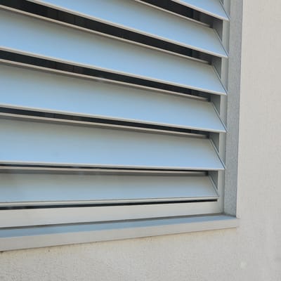 A close-up, eye-level shot shows a section of a building's exterior wall in Cuarte de Huerva, Spain. The wall is textured and painted a light gray or off-white. To the left, a ventilation louvre system is visible. The louvres are horizontal and made of a metallic material, appearing to be silver or light aluminum. They are angled downwards, suggesting they are designed to allow airflow while preventing rain from entering. The louvre system is set within a gray frame. The background through the louvres appears to be a light blue sky. The image is static and shows no people or activity.