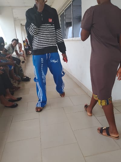 The scene is indoors on a tiled floor, possibly a waiting area or hallway, filled with people. In the foreground, two figures are prominent: a person in blue sweatpants with white stripes and the words "DON DOLLARA" on the leg, paired with a black and white striped top, is walking. Beside them, another person wears a brown dress with a patterned hem and black sandals. In the background, other individuals are sitting or standing, some engaged in conversation. The location is Umueshi, Nigeria.
