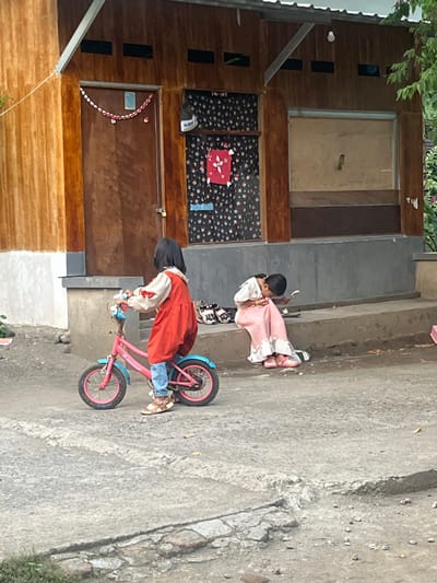 The scene depicts two young children in Desa Batu Layar, Indonesia, in front of a modest building. The foreground features an uneven, cracked concrete or asphalt surface.
To the left, a young child with dark, shoulder-length hair, wearing a white long-sleeved shirt under a red pinafore dress, blue jeans, and sandals, stands astride a small, pink bicycle with blue accents and training wheels. The child faces slightly away from the viewer towards the right.
To the right, another young child with dark hair, wearing a long, light pink dress with ruffled trim and pink open-toed shoes, sits on a grey concrete stoop. This child is leaning forward, holding a spoon, and appears to be eating from a small, dark bowl or plate positioned on the stoop. Behind this child, several pairs of sandals and slippers are neatly arranged on the stoop.
The background consists of a one-story building. Its upper half is clad in vertical wooden planks, while the lower half is a grey concrete base. The roof is corrugated metal, supported by metal beams. A wooden door, secured with a padlock, is visible on the left side of the building, adorned with a red and white chain. To the right of the door, a window is covered by a dark, floral-patterned curtain, with a red fabric item and a blue item hanging on it. Further right, another opening is boarded up with light-colored wood. A single bare light bulb hangs from the roof overhang above the boarded-up window. Green foliage is visible on the far right edge of the frame.