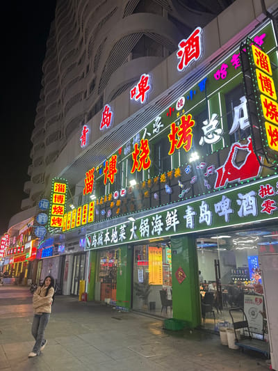 The scene depicts a vibrant commercial street at night in Huangdao District, China. A multi-story building dominates the background, featuring a residential or hotel section with a distinct wavy, patterned facade on its upper floors, unlit against a dark sky. The ground floor comprises a series of brightly lit commercial establishments, primarily a restaurant or bar with a prominent green facade.
The main establishment features extensive, illuminated signage in Chinese characters, predominantly in red, yellow, and white neon/LED lights. Key phrases visible include "青岛啤酒" (Qingdao Beer), "淄博烧烤" (Zibo Barbecue), "大锅海鲜" (Big Pot Seafood), and "酒" (Alcohol). Musical note symbols are also part of the signage design. Through large glass windows, the interior of the restaurant is visible, showing patrons seated at tables, along with some staff.
On the paved sidewalk in front of the establishments, a young East Asian female is positioned on the left side of the frame. She has long dark hair and is wearing a light-colored, possibly beige, long-sleeved top or jacket, light blue jeans, and white sneakers. She is leaning against a vertical support or light pole, with her arms crossed, looking directly towards the viewer.
To the left of the main establishment, more brightly lit signs in various colors extend down the street, indicating additional businesses. On the right side of the main storefront, several green plastic crates, two white buckets, and a grey hand truck are visible on the sidewalk. A red "Fu" character decoration is affixed to the glass door. The overall environment is brightly illuminated by the commercial signs, casting light onto the sidewalk.