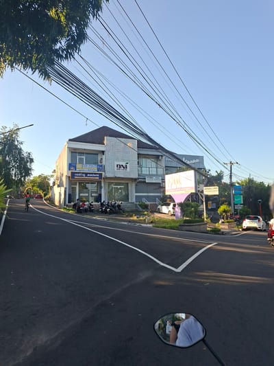 The image captures a street scene in Gianyar, Indonesia, likely on Jalan Gatot Subroto. A two-story commercial building with multiple businesses is prominently featured on the right side of the road. The building's facade displays several signs, including "DN SKIN CENTRE," "Piaggio Vespa," and "NOTARIS • PPAT." A smaller sign above a doorway reads "Pusat Belajar Coding AI & Robotika" and "coding academi." Several motorcycles are parked in front of the building.
On the left side of the road, a young person on a bicycle cycles away from the camera. Further down the road, a red car is visible. The street is paved asphalt with white lane markings. Overhead, a dense network of electrical wires crisscrosses the sky, casting shadows. The sky is clear and blue, indicating a sunny day. Lush green trees and foliage line the sides of the road, suggesting a tropical environment. In the foreground, a motorcycle's side mirror reflects the rider wearing a white shirt and a partial view of another person, indicating the photo is taken from a moving motorcycle. The overall scene depicts a typical urban street in Indonesia with commercial activity and transportation.