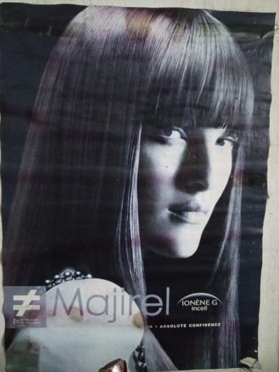 This image depicts a weathered advertisement poster affixed to a light-colored wall in the city of Keo Kouang, Laos. The poster features a female model, presented from the chest up, with her head turned slightly right and eyes directed forward. She has long, straight, dark hair with full bangs, showing subtle purple or reddish streaks. Her facial features include high cheekbones, a defined nose, and full lips, with a faint pink tone on her cheeks. A string of dark, spherical beads is visible around her neck/chest area. The poster prominently displays "Majirel" in white text, partially overlaying a soft, indistinct white circular object. Below this, smaller text reads "IONÈNE G incell" and "ABSOLUTE CONFIDENCE." To the lower left, a stylized symbol resembling a crossed-out hashtag is present with the text "ALL DIFFERENT EACH UNIQUE." The poster exhibits signs of age and wear, including creases, minor tears along its edges, and general discoloration or dirt accumulation. It is adhered to a plain, vertical surface, portions of which are visible surrounding the poster.