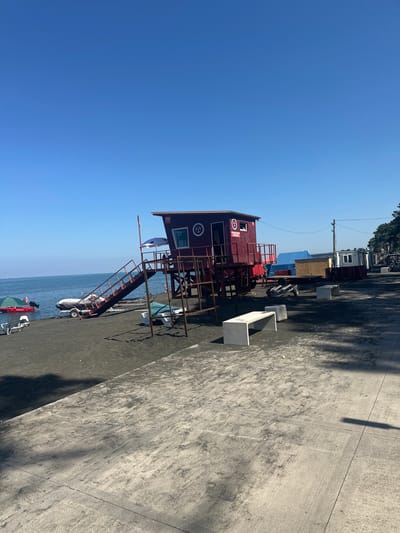 The image depicts a sunny day at a beach in Ureki, Georgia. In the foreground, a concrete promenade is visible with shadows cast by unseen objects. Beyond the promenade, dark sand covers the beach, on which several white benches and loungers are placed. To the left, a red emergency boat with a white canopy and the word "EMERGENCY" is docked near the water's edge. A trailer with a covered boat sits on the sand further back. The central focus of the image is a red lifeguard tower with a ramp leading up to it. The tower has a sign with "112" and other emblems. The calm blue sea stretches out to the horizon, meeting a clear blue sky. In the background, trees and several temporary structures are lined up along the shore. No people are clearly visible in the scene.