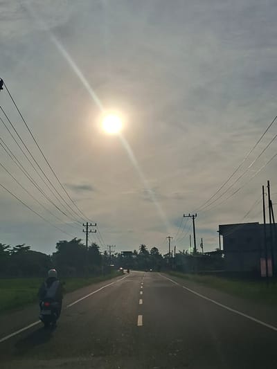 The image depicts a road scene in Bateiiliek, Indonesia. The sky is overcast with the sun prominently visible, casting a strong light and creating a lens flare effect that streaks across the frame. Power lines stretch across the upper portion of the image, supported by wooden utility poles. On the left side of the road, a person is riding a motorcycle, viewed from behind. The motorcycle has its taillight illuminated. The road itself is asphalt with white lane markings. To the right of the road, a dark, two-story building is visible. The surrounding environment includes scattered trees and foliage, with a hint of a green field on the left. The overall impression is that of a rural or semi-rural road on a bright, cloudy day.