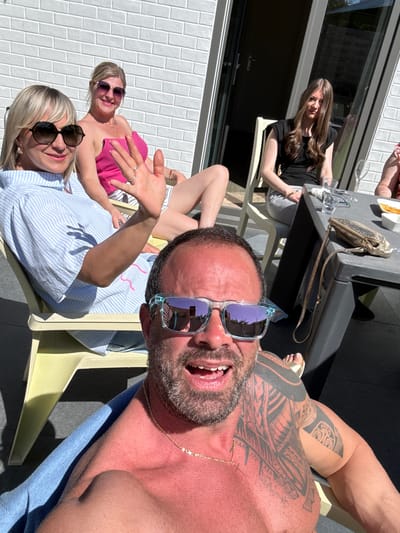 The image shows a group of people relaxing outdoors, possibly in Bredene, Belgium. A shirtless man with tattoos and sunglasses is prominently featured in the foreground. Three women are visible in the background: one in a black top, another in a pink top, and one in a blue and white striped shirt. They are seated on lawn chairs or loungers near a table with food and drinks. The setting includes a building with a white brick wall and a window. The scene appears sunny.