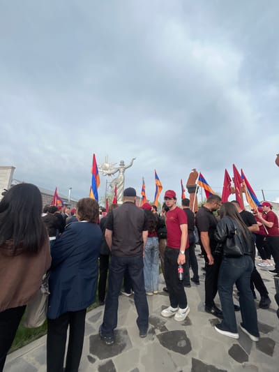 A photograph captures a gathering of people in Akhuryan, Armenia. They are clustered in a line near a prominent statue, each holding a flag displaying a tricolor pattern. The sky above is overcast. The ground is covered with stone pavers.