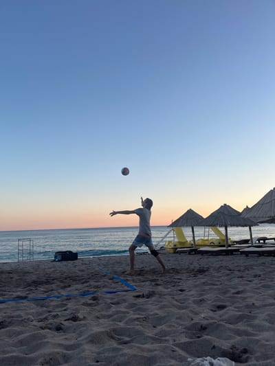 A lone male is captured in mid-action playing volleyball on a sandy beach in Miljevci, Montenegro. The sky displays a gradient of blues and soft oranges, indicating either sunrise or sunset. The man, wearing a gray t-shirt and blue shorts, is positioned with his arms extended, preparing to hit a volleyball that is suspended in the air above him. A blue volleyball net is partially visible on the sand in the foreground, delineating a court. In the background, the calm sea meets the horizon, with a few beach umbrellas and yellow paddle boats providing a backdrop. The sand is textured with footprints and small pebbles, and a few scattered items, including a black bag and a metal structure, are present on the beach.