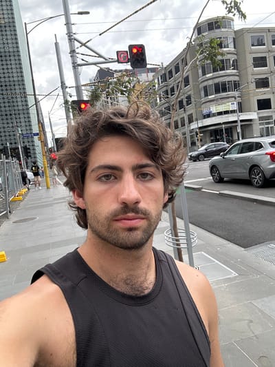 A male individual with brown, curly hair and a short beard is depicted from the mid-face to the upper chest, directly facing the camera. He is wearing a black sleeveless athletic top, and chest hair is visible.

The scene is an urban intersection in Melbourne, Australia, specifically at Victoria Street and King Street.

In the background, to the left, a modern high-rise building with a textured, patterned facade is partially visible. Construction barriers with yellow bases line the sidewalk, behind which two indistinct pedestrians are present. A street sign displaying "Victoria St" is mounted on a pole, near a black traffic light pole showing a red signal.

To the right, an older, multi-story building with a distinctive rounded corner facade and numerous windows occupies the mid-ground. Overhead wires, consistent with tram infrastructure, crisscross above the street. Two traffic lights are visible: one displays a red circular light, and the other a red 'no right turn' arrow. Two vehicles, a light grey SUV and a dark sedan, are on the road. A small tree with a metallic guard is present on the grey paved sidewalk. The sky is overcast.