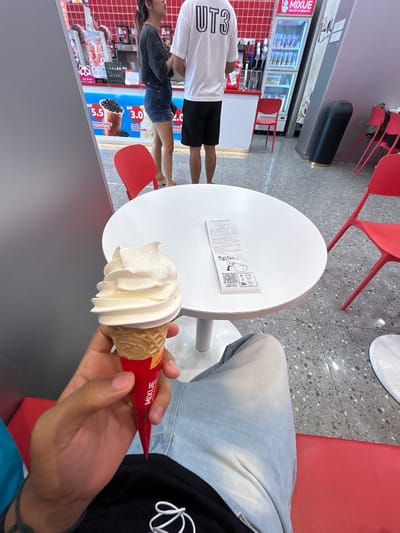 The image captures an indoor scene at a Mixue store, likely a cafe or dessert shop, characterized by its modern, clean aesthetic with grey speckled flooring and red and white furniture. In the foreground, a person's hand holds a creamy white soft-serve ice cream cone, encased in a red wrapper featuring the "MIXUE" logo and the word "Easy." This person is wearing light blue jeans over a dark long-sleeved shirt.
On a white round table beside them, a crumpled white paper or receipt is visible, displaying a QR code, the text "Fresh Fruit," and "ENJOY GIFTS." The receipt also indicates "Total products 2" and "BOMIXUE Local1257."
In the background, two other individuals are present. One person stands prominently, wearing a white t-shirt with "UT3" printed on the back and black shorts. Next to them, another person is partially visible, dressed in a dark top and denim shorts. They appear to be near the service counter or a display area.
The background features a refrigerator displaying beverages, a white counter, and various promotional signs characteristic of a Mixue establishment. These signs advertise "Brown Sugar Bubble Tea" for "RM 5.5," "Fresh Lemonade" for "RM 3.0," and "MIXUE Ice Cream" for "RM 2.0," using "RM" which denotes Malaysian Ringgit, aligning with the provided context of Subang Jaya City Council, Malaysia. Red chairs are scattered around, some stacked, and a black trash can is also visible. The overall lighting suggests it is daytime.