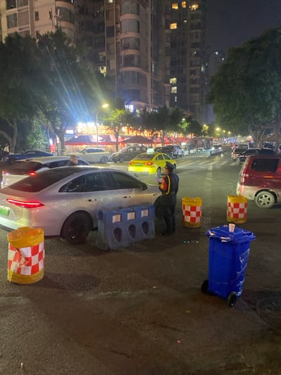This image depicts a nighttime urban street scene in 天宫殿街道, China. The scene is illuminated by numerous artificial light sources, including streetlights, vehicle lights, and lights from commercial establishments and residential buildings.
In the foreground and midground, a dark asphalt road surface is visible. A silver or light grey four-door sedan, possibly an electric vehicle indicated by a green license plate, is parked or stopped in the left midground, facing away from the camera. Adjacent to the passenger side of this vehicle is a blue plastic water-filled barrier with three recessed vertical segments. One yellow and white checkered cylindrical traffic barrier is in the left foreground, and two more are located in the right midground. A blue wheeled plastic trash bin with a white cup on its lid stands in the right foreground.
A single individual, dressed in a dark uniform with yellow or gold stripes on the sleeves and collar, stands facing right, between the silver sedan and the right-side traffic barriers. This person appears to be observing a small electronic device held in their hands.
Further down the street, multiple vehicles are visible, including a bright yellow taxi or ride-share car with a green license plate. A red minivan or SUV is partially visible on the far right. The street extends into the background, where more vehicles are present, indicating active traffic or parking.
The background features tall, multi-story buildings, likely residential or mixed-use, with numerous illuminated windows. Trees line the street, interspersed with streetlights. Below the buildings, several commercial establishments with brightly lit signs and awnings are visible, suggesting a busy commercial district. The sky is dark, consistent with nighttime.