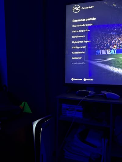 The scene depicts an indoor environment, specifically a room located in Chiapa de Corzo, Mexico, characterized by low ambient light and a predominant blue illumination. No individuals are present within the frame.
A large television screen, positioned in the upper right quadrant, displays a video game interface in Spanish. The screen shows a menu from "MC Carrera de DT" (Manager Career), with options including "Reanudar partido" (Resume match), "Dirección del equipo" (Team management), and "Configuración" (Settings). The background imagery on the screen depicts a football stadium with a crowd and a segment of a green pitch, marked with a "#FOOTBALL" hashtag.
Below the television, a dark-colored, two-tiered shelf unit is visible. The top shelf holds two white video game controllers, one of which is connected by a white cable. The bottom shelf contains a stack of horizontally arranged video game cases and a loose pile of white papers or documents.
To the immediate left of the shelf unit, a white PlayStation 5 console stands vertically. Its internal lighting emits a distinct white glow from its central vents, contrasting with the room's overall blue hue. Further to the left, partially obscured, is an object displaying a neon green surface. The room's walls are dark and uniform, reflecting the blue light from the television.