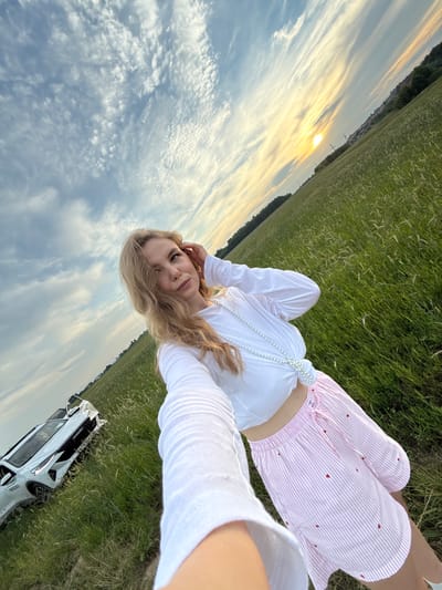 In the city of Сновицы, Russia, a young woman with blonde hair takes a selfie in a field of tall green grass, with a white SUV parked on her left. The sun is setting on the horizon, casting a warm glow on the scene. The woman is wearing a white long-sleeved top and a pink and white striped skirt with red heart patterns. She is also wearing a pearl necklace. The sky is filled with wispy clouds, and the overall atmosphere is serene and picturesque.