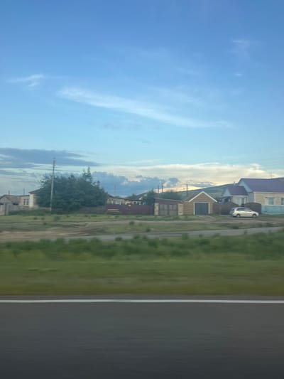 The image depicts a pastoral scene in Dzhirim, Russia. The orientation of the photograph is vertical. The sky, a soft blue with wispy white clouds, dominates the left side of the frame. In the middle ground, a row of houses with red-tiled roofs and wooden facades lines a dirt road. A silver car is parked in front of one of the houses. Green fields with some trees extend to the right, adjacent to a paved road with a white line down the center. The time of day appears to be late afternoon or early evening, given the soft light and the long shadows. The weather seems pleasant, with clear skies and fair visibility. The overall atmosphere is tranquil and evokes a sense of rural life. There is no visible text in the image.