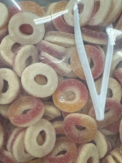 A close-up, top-down shot captures a pile of ring-shaped candies, possibly gummy rings, submerged in a clear plastic bag. The candies are predominantly a pale yellow or cream color, with distinct pink or reddish bands encircling their outer edges. A scattering of sugar crystals is visible on the surfaces of the candies, giving them a slightly frosted appearance.
Two white plastic tongs are partially visible, positioned in the upper-middle section of the frame, with their tips grasping some of the candies. The tongs appear to be either for serving or holding the candies within the bag. The lighting suggests an indoor setting, with some reflections on the plastic surface indicating overhead light sources. The overall impression is of a sweet treat assortment, likely a product for sale or consumption. No people, text, specific location cues, time of day, or weather conditions are discernible in the image.