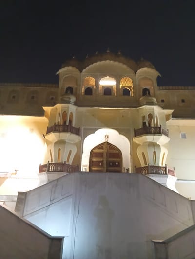This night photograph captures a grand, illuminated building, likely a historical palace or a significant architectural structure, against a dark sky. The scene is devoid of people, focusing entirely on the edifice.

The building displays elaborate Indian, specifically Rajasthani, architectural styles, aligning with the provided context of Jaipur. It is constructed from a light-colored material, possibly stone or stucco, which glows warmly under artificial lighting.

A prominent feature in the center of the image is a large, ornate wooden door with intricate details and brass accents, set within a grand arched entrance. Above and surrounding this entrance, the building rises several stories, showcasing multiple arched windows, some with delicate latticework (jalis), through which warm, golden light emanates. Other windows are open arches revealing internal illumination. Several balconies with finely crafted wooden railings project from the facade, adding depth and shadow to the structure. Dome-like elements or turrets crown the upper sections of the building.

The entire building is bathed in a bright, warm yellow light, creating a stark and beautiful contrast against the dark, clear night sky that forms the background. The intense illumination highlights the intricate carvings, arches, and architectural details. To the right of the main structure, a simpler, lighter-colored wall of another building or a closer architectural element is visible, slightly obscuring part of the main facade. A very dark area in the lower-left suggests a possible reflection in a water body or a very dark foreground.

No specific activity or event is occurring, and there is no visible text in the image. The overall impression is one of serene nighttime beauty, showcasing the architectural grandeur of a significant landmark.