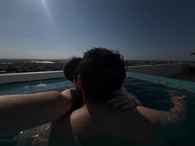 A person is submerged in a rooftop swimming pool in Tampico, Mexico. The person's head is above the water, and their face is obscured by hair and dark-colored goggles. They are looking out over the city from the rooftop pool, resting their chin on their folded hands. Sunlight is reflected off the city skyline. The pool's tiled edge is in the foreground.