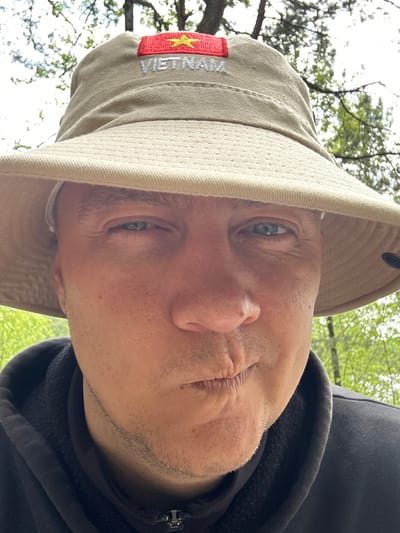 A close-up, rotated portrait of a middle-aged man wearing a khaki-colored sun hat with a Vietnamese flag and the word "VIETNAM" embroidered on the side. He is also wearing a dark hooded sweatshirt. The background is blurred, showing trees and foliage, suggesting an outdoor location. The man has light skin, blue eyes, and a stubbled beard. He is looking at the camera with a slightly puckered expression. The photo seems to be taken from a low angle, looking upwards. The scene appears to be in a natural setting, possibly a park or wooded area, but no specific details indicate it is Юзуфоўскі сельскі Савет, Belarus.