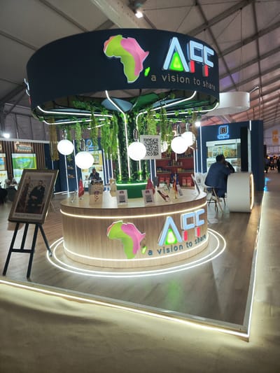 This media file presents an indoor view of an exhibition booth, likely situated at a trade show or convention in Al Machouar – Stinia, Morocco. The central feature is a circular booth with a wooden-textured counter and a suspended canopy, both adorned with white LED strip lighting. The booth's branding, "AFF a vision to share," is prominently displayed on both the counter and canopy, accompanied by a stylized, multi-colored map of Africa.
A tree-like structure emerges from the center of the counter, supporting the canopy and featuring hanging artificial green foliage and several spherical white pendant lights. Additional greenery descends from the canopy, enhancing a natural aesthetic. The counter holds various items, including small flags (one appearing to be Moroccan), brochures, and stands with QR codes. A larger QR code is visible hanging from the tree structure.
Several people are present, indicating ongoing activity. A man in a suit is seated at the central booth's counter, facing away, while another man is at a nearby blue-themed booth branded "BAGAN," which features product displays. Two other men are seated at a table to the left.
To the left of the main booth, a large framed portrait of a man, consistent with King Mohammed VI of Morocco, is displayed on an easel. This, along with a Moroccan flag on the counter, provides strong location cues. The setting includes a large, tent-like ceiling and other exhibition stands in the background. The scene is well-lit, conveying a professional and engaging environment. Visible text includes "AFF a vision to share," "BAGAN," and an unidentifiable partial "7."