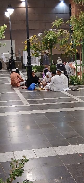 The image depicts an outdoor, paved plaza or walkway at night in Medina, Saudi Arabia. The ground consists of dark grey tiles with lighter grey accent stripes. In the central foreground, a group of approximately five to six individuals are seated on the paved surface, arranged in a rough circle, sharing a communal meal laid out on a light-colored mat. Visible items include plates of food and a plastic water bottle. The seated individuals wear traditional attire, including men in white thobes and head coverings, a woman in a black abaya and headscarf, a person in a light-colored abaya and white head covering, and another person in an orange-brown thobe. A blue plastic bag is placed near the group.

In the mid-ground, a man wearing a light-colored jacket and a white baseball cap walks towards the left. Scattered in the background are additional individuals, some seated on the ground or on low structures. The environment includes multiple tall, black decorative lamp posts with illuminated white lights. Small trees and bushes with green and some yellowish foliage are planted within defined areas. A large building with light-colored, block-patterned walls, illuminated by overhead lights, forms the backdrop. White utility structures are visible behind the trees.