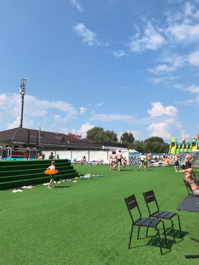 The image depicts an outdoor scene at a water park in Gamovo, Russia. The foreground features a vibrant green artificial turf area with several pairs of shoes scattered on the ground. A young boy, wearing green shorts, walks across the turf holding an orange inflatable ring. In the mid-ground, several people are visible, some walking and others relaxing on lounge chairs. A swimming pool is to the left, with people in and around it. Behind the pool, a building with a brown roof is present, featuring the red illuminated sign "Weekend" on its facade. Further in the background, a large inflatable water slide structure is set up, with people interacting with it. The sky is a clear blue, dotted with fluffy white clouds. A tall cell tower stands to the left of the frame. Two dark metal chairs and a table are placed on the artificial turf in the lower right corner.