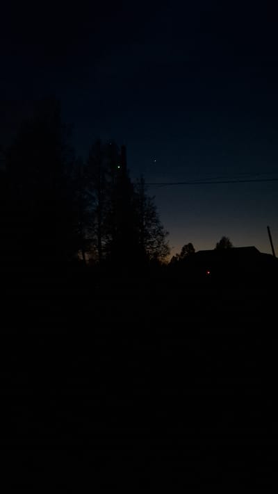 A nighttime scene in Шешурга, Russia, features a dark sky transitioning from deep blue to a lighter hue near the horizon. Silhouetted trees dominate the foreground, with a structure or building looming to the right. Two points of light are visible: a green light amidst the trees, and a single red light emanating from the structure. Power lines stretch across the frame. The scene is dominated by darkness.
