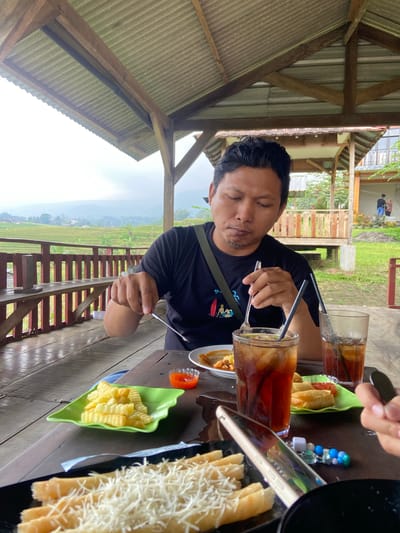 The photo shows a man sitting at a wooden table in an open-air structure with a corrugated metal roof. He wears a black t-shirt with a printed design and has a dark complexion. He is stirring a tall glass of dark liquid, likely iced tea, with a spoon. On the table, there are several plates with food, including French fries, spring rolls, and cheese sticks. Beside the plates are small dishes of what appears to be chili sauce. There are also glasses of iced tea. A smartphone is visible near the edge of the table. Outside the structure, a view of rice terraces and distant hills is visible, suggesting a rural or agricultural setting. The location is likely Penanggungan, Indonesia, judging by the type of scenery and structure shown. Other people are partially visible in the background and at the edge of the table.