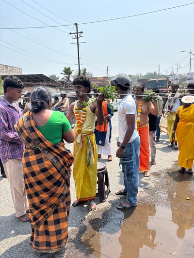 A group of individuals are participating in what appears to be a religious procession or ceremony on a street in Ambernath, India. The street is somewhat wet and reflects the sunlight. Participants are adorned in traditional Indian clothing, including sarees, dhotis, and flower garlands. Some are barefoot.

One person carries a small arrangement of green branches. A few individuals are wearing headgear, and there's a chair present in the procession's midst. Power lines and utility poles are visible above, indicating an urban setting. Various vehicles, including a truck, are also present in the background, suggesting the procession is taking place on a road with vehicular traffic.