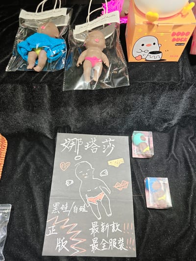 In the city of Gongshu District, China, a display table is covered with a black velvet cloth. On the table, there are several items for sale, including two small baby dolls in plastic packaging, a "Dodo Duck" night light in an orange box, and a black cardboard sign with drawings and text.
The baby dolls are approximately 4-6 inches in length. One doll is wearing a blue knitted dress and yellow underwear, while the other is wearing pink underwear. The dolls have dark skin and pacifiers in their mouths. The "Dodo Duck" night light features a cartoon duckling sitting on a pudding.
The black cardboard sign has hand-drawn illustrations of a baby, a heart, underwear, and lips. The text on the sign appears to be in Chinese and includes characters like "娜塔莎" (Natasha) and "黑娃/白娃" (black doll/white doll). There are also phrases that translate to "new style!!!" and "full clothing!!!". Two small clear plastic boxes, each containing a baby doll in a different outfit, are placed to the right of the sign. The overall scene suggests a market stall or retail display.