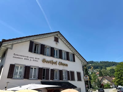 The image shows a white building with a gabled roof, inscribed with "Tavernenrecht anno 1640" and "renoviert 1992 J. Sidler". Above a row of windows, a golden sign reads "Gasthof Löwen". The building is located in Wald (ZH), Switzerland. The foreground shows outdoor seating with white umbrellas and tables. In the background, a street curves through a village with traditional Swiss architecture, surrounded by lush green hills and trees. A few cars are parked and driving on the street. The sky is clear and blue with a faint contrail from an airplane.