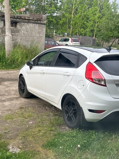 A white Ford Fiesta is parked on a dirt and gravel lot next to a dilapidated brick building. The car is angled, with its rear tire on the right side of the image partially submerged in tall green grass. The car has black rims and a visible license plate holder, but the license plate itself is unreadable. The building appears to be abandoned or under construction, with exposed brick and a rusty metal structure on top. Other cars are visible in the background, including a red minivan and another white SUV. The scene is outdoors, with trees and greenery surrounding the area. The sky is overcast, suggesting a cloudy day. The overall impression is one of disuse and neglect, with the car being the most prominent and well-maintained object in the frame. There is no visible text in the image.