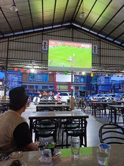 The image depicts an outdoor seating area of a restaurant or cafe in Bireuen, Indonesia, where patrons are watching a soccer match on a large screen. The screen displays the FIFA World Cup 2026 logo, indicating the event being broadcast. The seating area is covered by a corrugated metal roof supported by metal beams. Several tables and chairs are arranged for diners. A person in the foreground, wearing a black beanie and a light-colored shirt, is seated and looking towards the screen. In the background, there are parked motorcycles and cars, as well as buildings with various advertisements and banners. Flags of different nations are hung across the area, likely to represent the countries participating in the soccer tournament. The lighting suggests it is evening or nighttime.