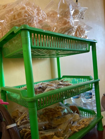 The image shows a green, three-tiered plastic shelving unit displaying various packaged snacks. The top shelf holds large, clear plastic bags filled with pale, coiled snacks, possibly crackers or krupuk. The second shelf contains clear plastic bags filled with smaller, irregularly shaped, golden-brown snacks. The bottom shelf also holds clear plastic bags of similar golden-brown snacks, with two bottles of "Prima" bottled water visible behind them. The shelving unit is placed against a textured, yellow wall. To the left of the unit, a portion of a dark brown bag or strap with a white tie is visible. The scene appears to be indoors, likely a small shop or stall, given the presentation of goods. The context suggests a location in Gianyar, Indonesia, where such snacks are commonly sold.
