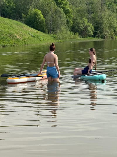 Two women are wading in a body of water, each with a paddleboard. The woman closest to the foreground is on the left side of the frame, facing forward and to the right. She is wearing a blue bikini top and matching blue shorts. Her brown hair is pulled back into a bun. She is leaning down towards the paddleboard, with her right hand touching it. A black paddle lies in the water near the paddleboard. The woman on the right side of the frame is wearing a brown bikini top and blue shorts. Her brown hair is styled in a long braid. She is kneeling on the paddleboard, bending forward and looking down into the water. The water is reflective, with a rippled surface. Lush green vegetation and trees are present along the bank on the left side of the frame. Based on the prompt instructions, the location is Novosinkovo, Russia.