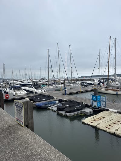 The image shows a marina in Poole, United Kingdom. The sky is overcast. Several sailboats and motorboats are docked at wooden piers. Some jet skis are covered with black tarps and parked on a floating platform. A yellow sign indicating "PRIVATE LAND AUTHORISED USERS ONLY PARKING" is attached to a wooden post on the left side of the image. There are no people clearly visible in the frame. The water is dark and calm.
