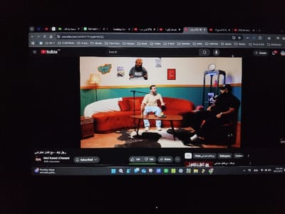 The image shows a laptop screen displaying a YouTube video. The video is a talk show featuring two men sitting on a red couch. The man on the left is wearing a white shirt and blue jeans, while the man on the right is wearing a black shirt and black pants. Both men are facing the camera. There is a coffee table in front of them with a microphone and a glass of water. The wall behind them is decorated with a blue and teal paint scheme with various posters and stickers. The video is titled "مع كمال - حلقة الخميس" (With Kamal - Thursday episode). The YouTube channel is named "MC3 Kamel A7erami" and has 43K subscribers. The laptop is located in the city of Al Zahraa WA Masaken Al Helmeyah, Egypt, based on coordinates (latitude: 30.12, longitude: 31.339).  The scene is illuminated by bright overhead lighting.  There is a dark, black background surrounding the laptop, suggesting it may be placed on a table or desk in a darkened room.  There are several other windows open on the laptop screen but none are visible.  The image appears to be captured from a slightly elevated angle, as though the viewer is looking down on the laptop from above.  The screen is bright and clear, suggesting the laptop is in active use.  The image is taken during the day as there is natural light visible in the window at the top of the laptop screen.