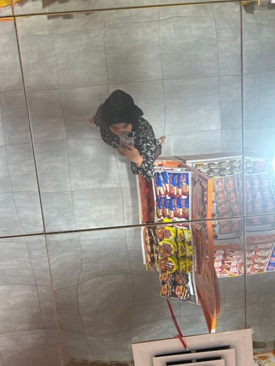 This media file captures a reflection of a person in a mirrored surface, likely a wall or a display. The person is wearing a black hijab and a patterned black and white dress, and is holding a smartphone, taking a selfie. They are standing in what appears to be an aisle in a store, as indicated by the reflection of shelves stocked with various food products, predominantly instant noodles and snacks.

The setting is a retail environment, with tiled flooring visible in the reflection. The lighting suggests an indoor setting, possibly a supermarket or convenience store. The quality of the reflection is distorted, characteristic of a warped mirror or a curved surface, creating a slightly disorienting effect.

The visible text on the products is in a language that appears to be Indonesian or Malay, with labels such as "INDOMIE" and other product names that are partially legible due to the reflection. The overall impression is of a casual moment captured in a public place, with the person interacting with their phone in a mirrored environment. The time of day and weather are not discernible from the image. The emotion conveyed is neutral, as is typical for a selfie in a retail setting.